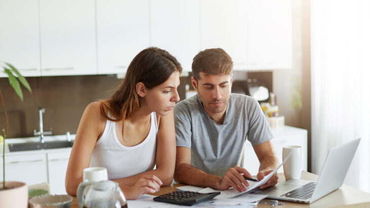 Pareja joven revisando cuentas y organizando sus finanzas en pareja en la mesa de su hogar, con computadora, facturas y calculadora.